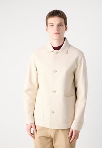 Light beige jacket with a collar, featuring three front buttons, two front pockets, and a smooth texture. Worn over a burgundy shirt.