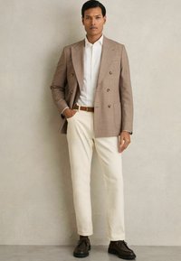 Sacou maro cu carouri, cu două rânduri de nasturi, cămașă albă, pantaloni crem, curea din piele maro și pantofi maro închis. Texturi netede în întreaga compoziție.