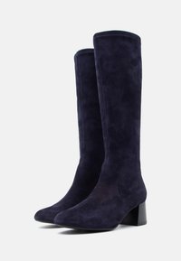 Egy pár magas, sötét tengerészkék velúr csizma hegyes orral és alacsony blokk sarkakkal egy sima, világos háttér előtt.