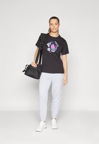 Tricou negru cu un print colorat, pantaloni de trening gri deschis și adidași albi. Ținând o geantă sport neagră, stând pe un fundal alb.