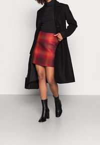 Una donna posa con sicurezza indossando un cappotto nero, un dolcevita e una mini gonna a quadri rossi, abbinati a stivali neri con tacco alla caviglia, su uno sfondo neutro.