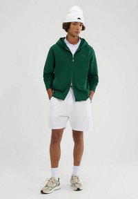 Sudadera verde con cremallera, polo blanco, pantalones cortos blancos y zapatillas multicolores. El modelo lleva un gorro blanco. Texturas suaves, ajuste relajado.