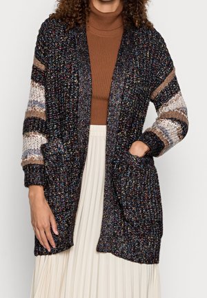 Femme portant un cardigan en maille épaisse multicolore avec des manches rayées, un col roulé marron et une jupe plissée blanche, main dans la poche.