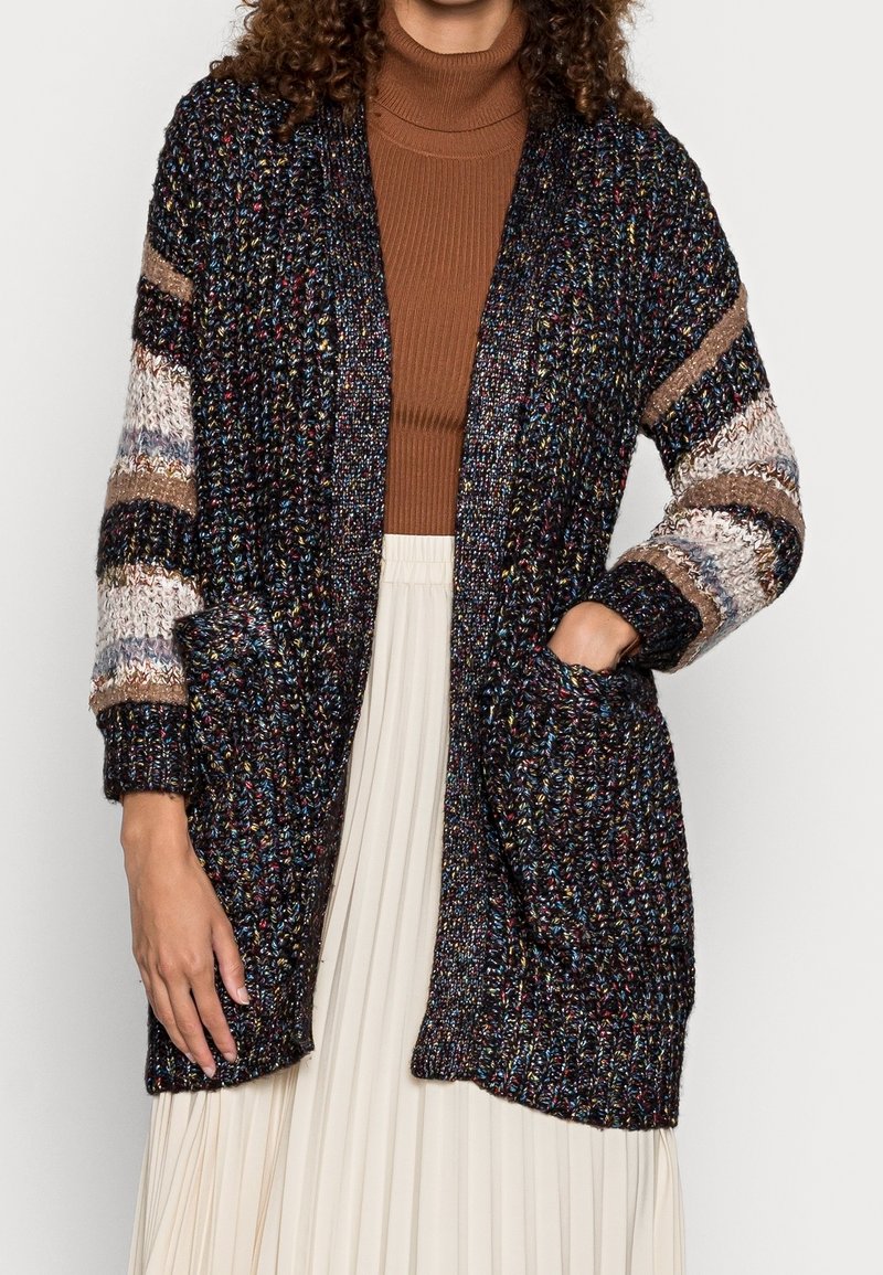 Femme portant un cardigan en maille épaisse multicolore avec des manches rayées, un col roulé marron et une jupe plissée blanche, main dans la poche.