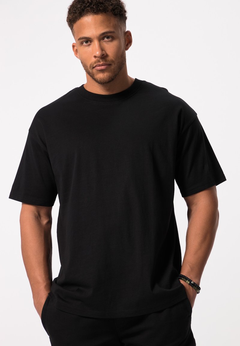Man met kort krullend haar, draagt een effen zwarte oversized korte mouw T-shirt, handen gedeeltelijk in de zakken, neutrale gezichtsuitdrukking, witte achtergrond.