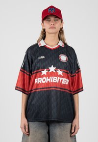 Mujer que lleva una camiseta deportiva negra y roja de gran tamaño con la palabra “PROHIBITED” y tres estrellas blancas, una gorra roja con un logo azul y pendientes de aro.