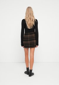 Schwarzes Langarmoberteil kombiniert mit einem braunen karierten Minirock, der schwarze Akzente und strukturierte Stoffe aufweist. Stilvoll ergänzt mit schwarzem Schuhwerk und Socken.