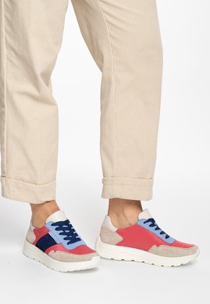 Colorful sneakers featuring red, blue, and beige panels, with a textured suede finish and blue laces, set against beige corduroy pants.