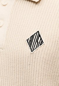 Κρεμ πλεκτό πουλόβερ με λευκά κουμπιά και ένα μικρό ασπρόμαυρο κεντητό σχέδιο με διαγώνιες ρίγες στο στήθος.