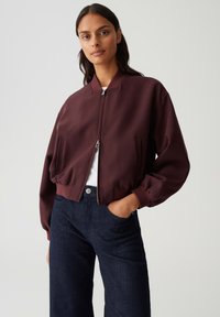 Giacca bomber bordeaux con zip frontale, polsini a costine e tasche laterali, abbinata a pantaloni in denim scuro, su uno sfondo neutro.
