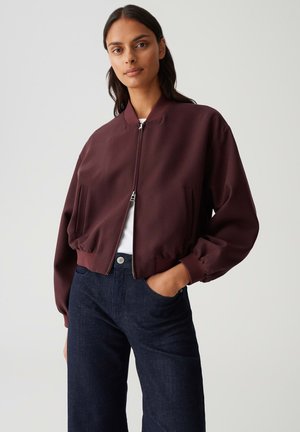 Burgundy bomber jacket with zip front, ribbed cuffs, and side pockets, paired with dark denim pants, against a plain backdrop.