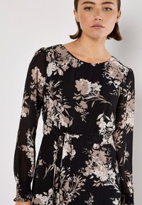 Blumenlangarmkleid mit schwarzem Hintergrund und beigen sowie cremefarbenen Blumen, Rundhalsausschnitt und einem Bindegürtel in der Taille. Glatte Textur.