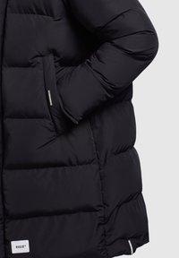 Fekete puffatjak, steppelt mintázattal, oldalsó zsebbel és egy kis márkacimkével az alján, sima szintetikus anyagból készült.