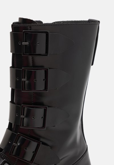 Black ankle boot made of glossy material, featuring three wide straps with rectangular buckles and a seamless rounded shape.