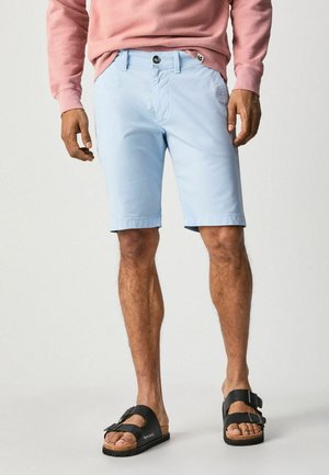Shorts bleu clair avec un bouton et une fermeture éclair, poches latérales et ourlets roulés, associés à des sandales en cuir noires avec un détail de boucle.
