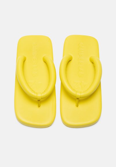 Des tongs jaunes avec une texture lisse, dotées d'un design à large bride et d'une semelle plate. Le nom de la marque est embossé sur la surface supérieure.