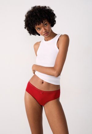 Slip da donna rossi con finitura a trama, bordatura in pizzo e piccolo fiocco come dettaglio. Abbinati a un top aderente bianco.