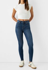 Jeans de denim azul de tiro alto con corte ajustado, que presentan bolsillos clásicos, detalles de costura y una textura suave. Combinados con un top corto blanco ajustado y zapatillas.