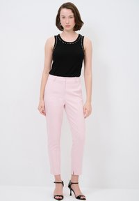 Zwarte mouwloze top met studs bij de halslijn; lichtroze op maat gemaakte broek; zwarte hoge sandalen; minimale accessoires, staande tegen een neutrale achtergrond.