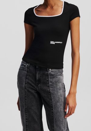 Kobieta ubrana w czarny dopasowany top z krótkim rękawem z białym obszyciem i napisem „KARL LAGERFELD JEANS”, w połączeniu z wysokim stanem i wyblakłymi czarnymi dżinsami.