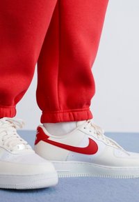 Sneakers Nike Air Force 1 bianchi con dettagli rossi, abbinate a pantaloni della tuta rossi con bordo elastico, poggiati su una superficie blu.