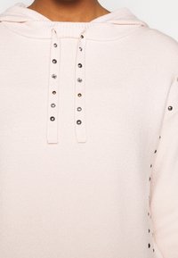 Ljusrosa huvtröja med metallnitar som dekorerar snoddarna och som är utspridda längs höger ärm.