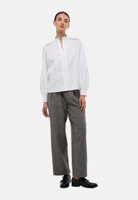 Blouse en coton blanc avec fermeture à boutons et plis subtils sur le devant, associée à un pantalon en pied-de-poule gris à jambes larges et des mocassins noirs.