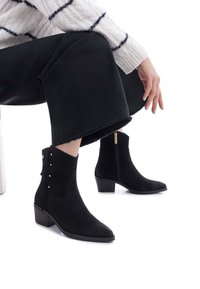 Botines de ante negro con puntera afilada, tacón bajo en bloque, cremallera dorada y tachuelas decorativas en el lateral. Se llevan con pantalones negros recortados.