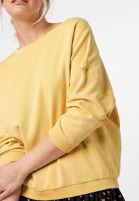Sudadera amarilla hecha de tela suave, con un corte holgado, cuello ancho y puños acanalados. Textura lisa con detalles de costura mínimos.