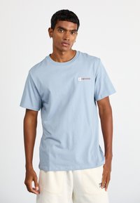 Lys blå bomulds T-shirt med rund hals, korte ærmer og et lille rødt og hvidt Converse-logo på venstre bryst.