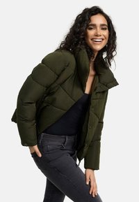 Dark green puffer jacket with a high collar, quilted design, and snap fasteners. Worn over a black top with slim-fit gray jeans.