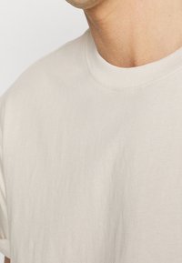 Gros plan d'une personne portant un t-shirt uni beige clair à col rond, montrant le cou et le haut de la poitrine.