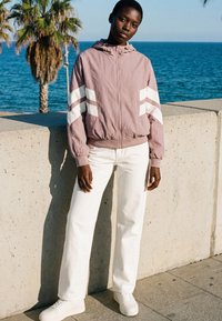 Femeie purtând o jachetă roz prăfuit cu dungi albe, pantaloni albi și adidași se sprijină de un perete de beton lângă mare, cu palmieri în apropiere.