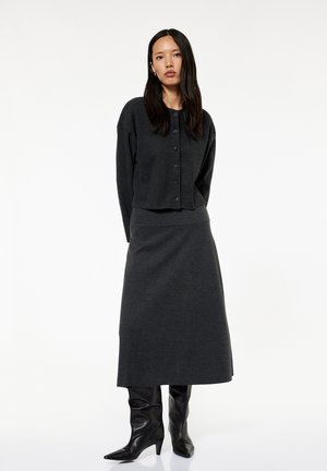 Kobieta o długich ciemnych włosach, ubrana w ciemnoszary rozpinany kardigan, dopasowaną długą spódnicę i czarne botki na obcasie, stojąca na białym tle.