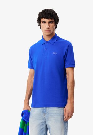 Polo en coton bleu à manches courtes, avec un plastron à trois boutons et un logo brodé sur la poitrine. Porté avec un jean bleu clair.