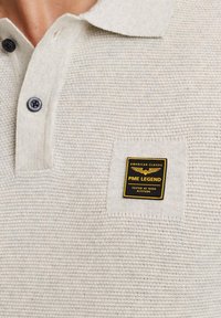 Helles graues Polohemd aus strukturiertem Stoff. Verfügt über einen weichen Kragen, zwei schwarze Knöpfe und ein quadratisches PME Legend Etikett auf der Brust.