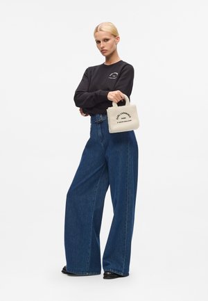 Black sweatshirt with circular logo, high-waisted blue denim wide-leg pants, and small cream fabric handbag with printed text.