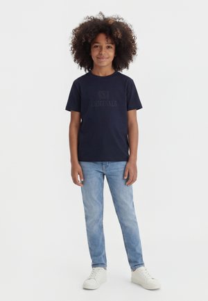 Enfant aux cheveux bouclés portant un t-shirt bleu marine avec le texte "Originals", un jean bleu clair et des baskets blanches, debout devant un fond blanc.
