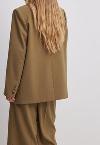 Marynarka w oliwkowym kolorze, wykonana z sztywnego materiału, w luźnym kroju, z zapięciem na jeden guzik i teksturowanymi guzikami, w zestawie z pasującymi spodniami.