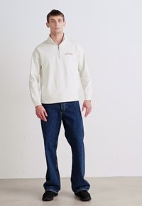 Suéter crema de peso medio con cuello con cremallera, que presenta un pequeño logo bordado. Se lleva con unos vaqueros azul de corte holgado y zapatos negros.