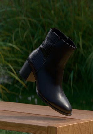 Bottine en cuir noir avec talon bloc et détail de texture crocodile, positionnée en l'air au-dessus d'une surface en bois en extérieur.