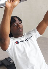 Camiseta de algodón blanca con el logo negro de "Champion", utilizada por una persona levantando pesas en un gimnasio; textura suave y corte informal.