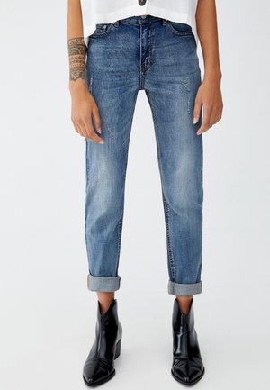 Jean en denim bleu clair avec une coupe décontractée, des ourlets roulés, un léger délavage et des zones d'effritement. Associé à des bottines noires.