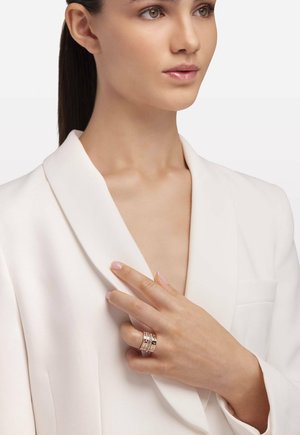 Weißer Blazer mit einem Schalkragen, getragen über einem V-Ausschnitt-Oberteil. Eine Hand präsentiert einen Mehrbandring mit abwechselnd silbernen und goldenen Tönen.
