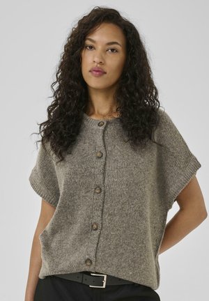 Frau mit lockigem dunklem Haar, die einen kurzärmeligen grauen Knopfstrickpullover und schwarze Hosen mit einem schwarzen Gürtel vor einem schlichten Hintergrund trägt.