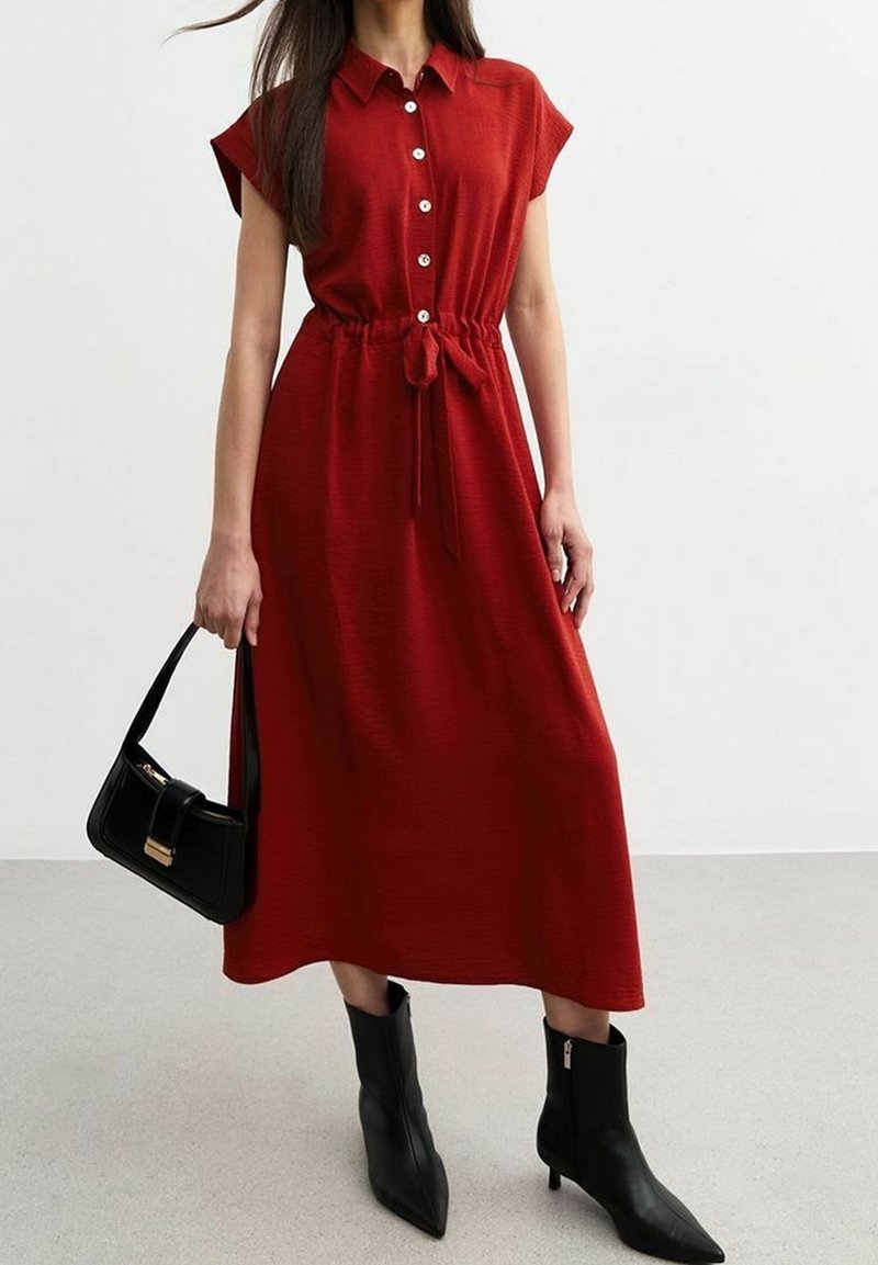 Robe rouge à boutons avec manches courtes, taille cintrée et détail avec lien. Associée à des bottines à talons noires et un sac à main noir structuré.