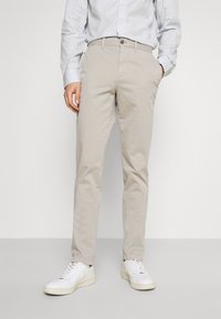 Beige Chinos in Slim-Fit aus Baumwolle, mit einem Reißverschluss und Knopfverschluss. Kombiniert mit weißen Sneakers und einem blau gestreiften Hemd.