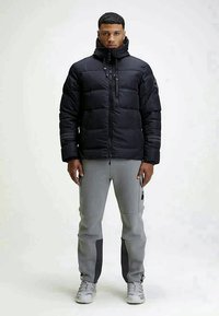 Schwarze Puffy-Jacke mit hohem Kragen, Reißverschluss und sichtbaren Nähten. Kombiniert mit grauen Hosen, die seitliche Taschen und kontrastierende Akzente aufweisen.