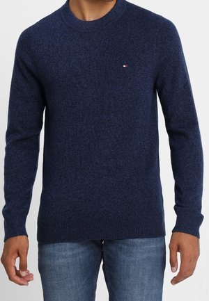 Man wearing a navy blue crewneck sweater with a small logo on the chest and blue jeans, standing against a plain background.