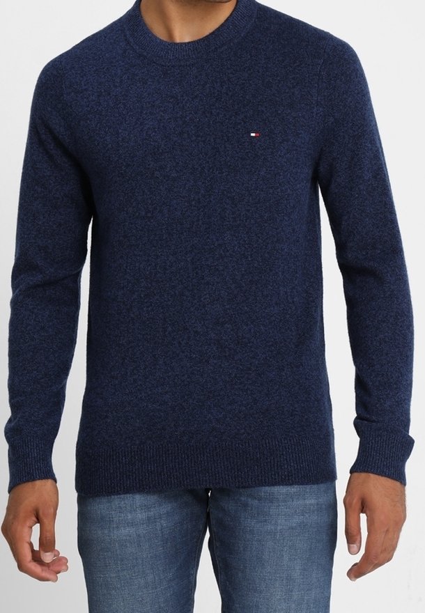 Homme portant un pull bleu marine à col rond avec un petit logo sur la poitrine et un jean bleu, debout devant un fond uni.